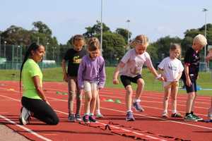 Sportspark instructor kneeling on athletics track while group of children run single file through athletics ladder.
