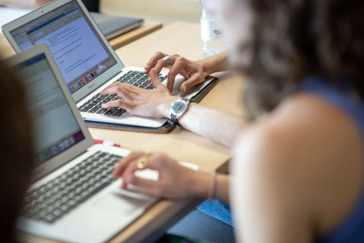 People typing on laptops at a desk, focused on work. A water bottle and papers are visible on the table.