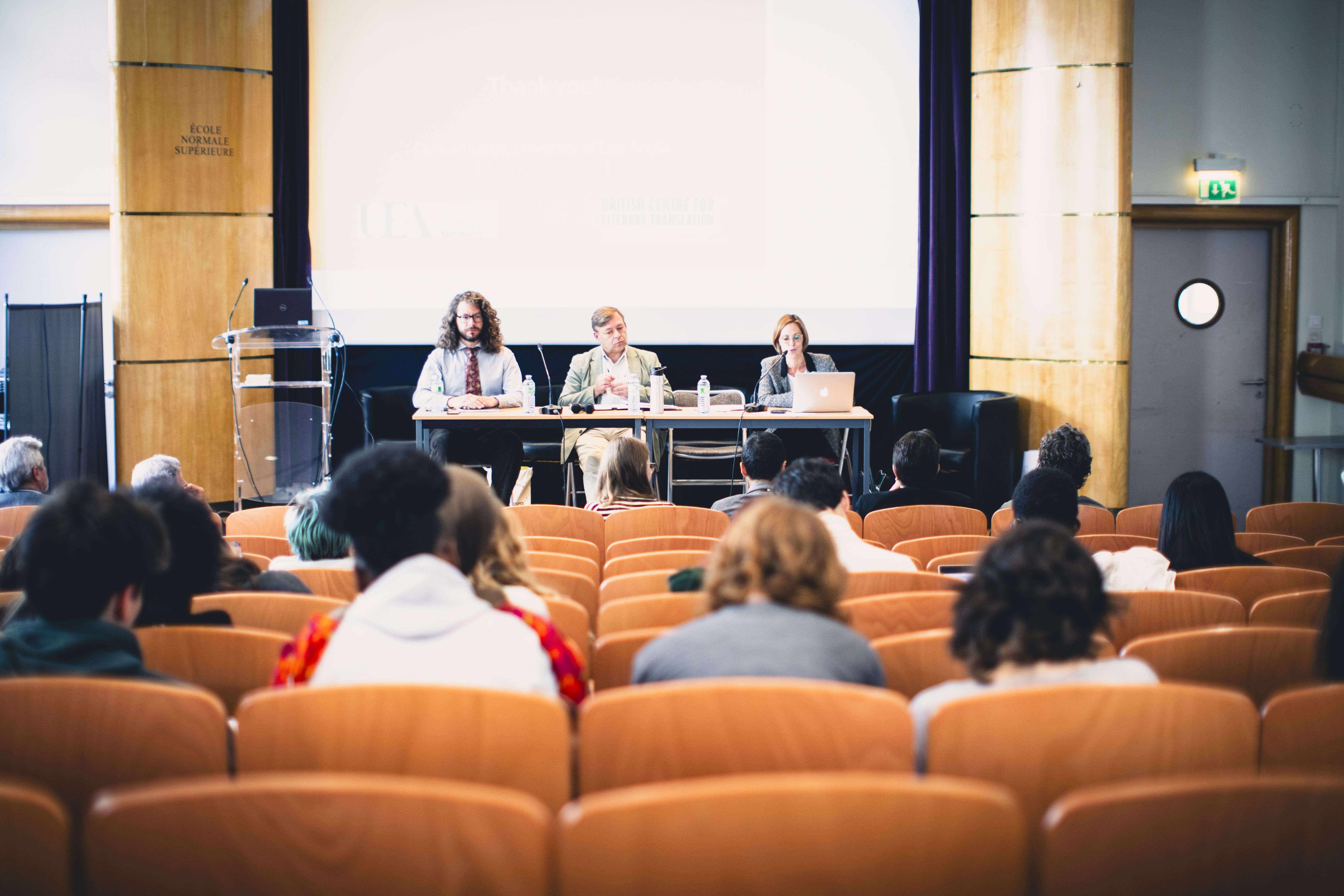 An audience in a lecture hall watches a panel of three speakers on stage during a presentation.