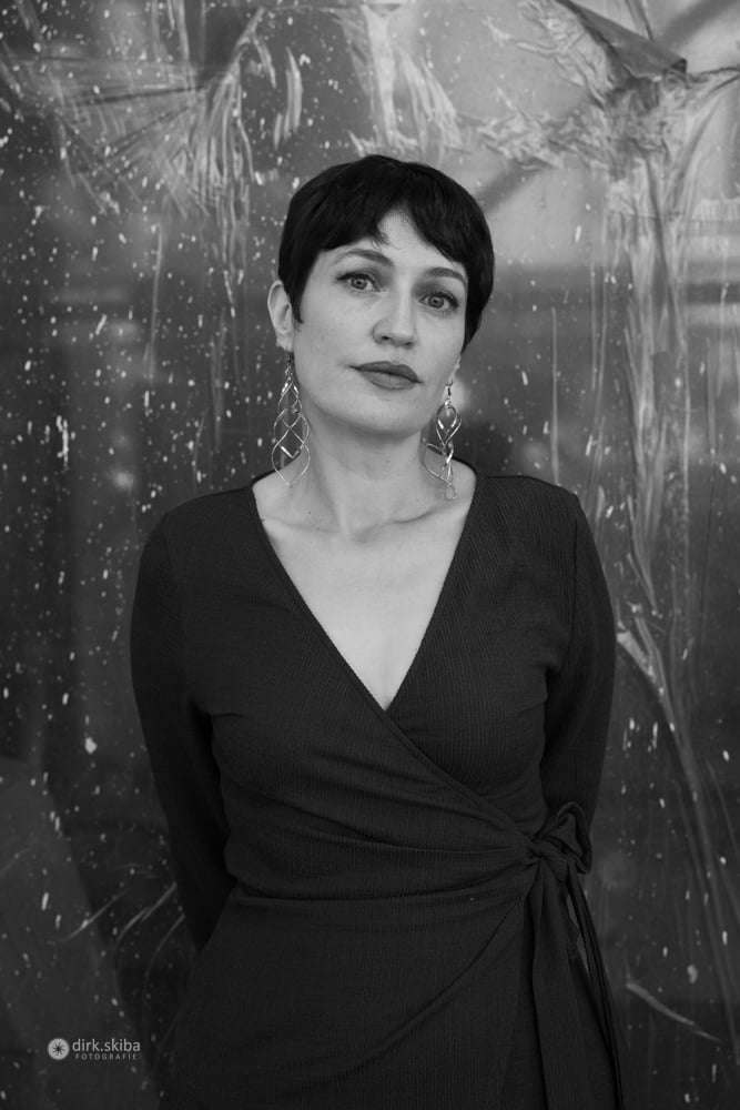 A black and white portrait of a woman with short dark hair and large, dangling earrings. She wears a dark v-neck wrap dress and looks at the camera.