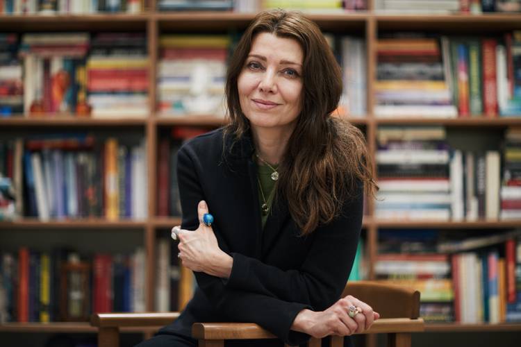 Elif Shafak has long dark hair. She is wearing a black blazer and sitting cross armed infront of a book shelf