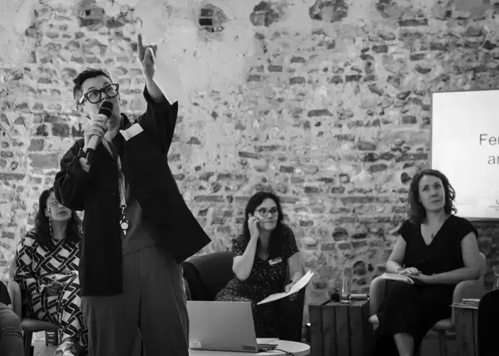 A person speaks into a microphone, gesturing upward, while three others sit, listening, in a room with a stone wall background.