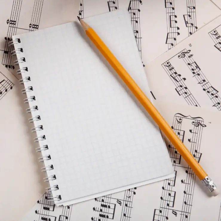 A blank spiral notebook and a yellow pencil rest on scattered pages of sheet music.