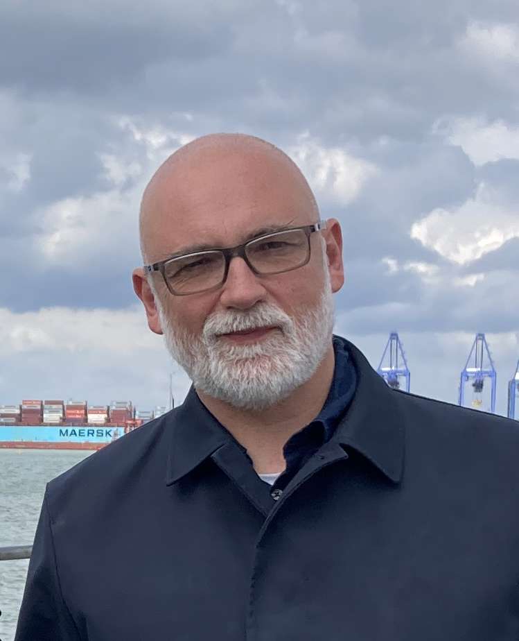Tom has  glasses and is wearing a dark jacket stands outdoors with a cargo ship and cranes in the cloudy background.