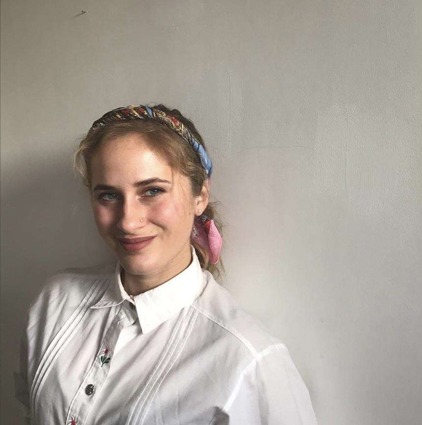 A smiling young woman with a colorful scarf in her blonde hair wears a white collared shirt with floral embroidery against a plain wall.