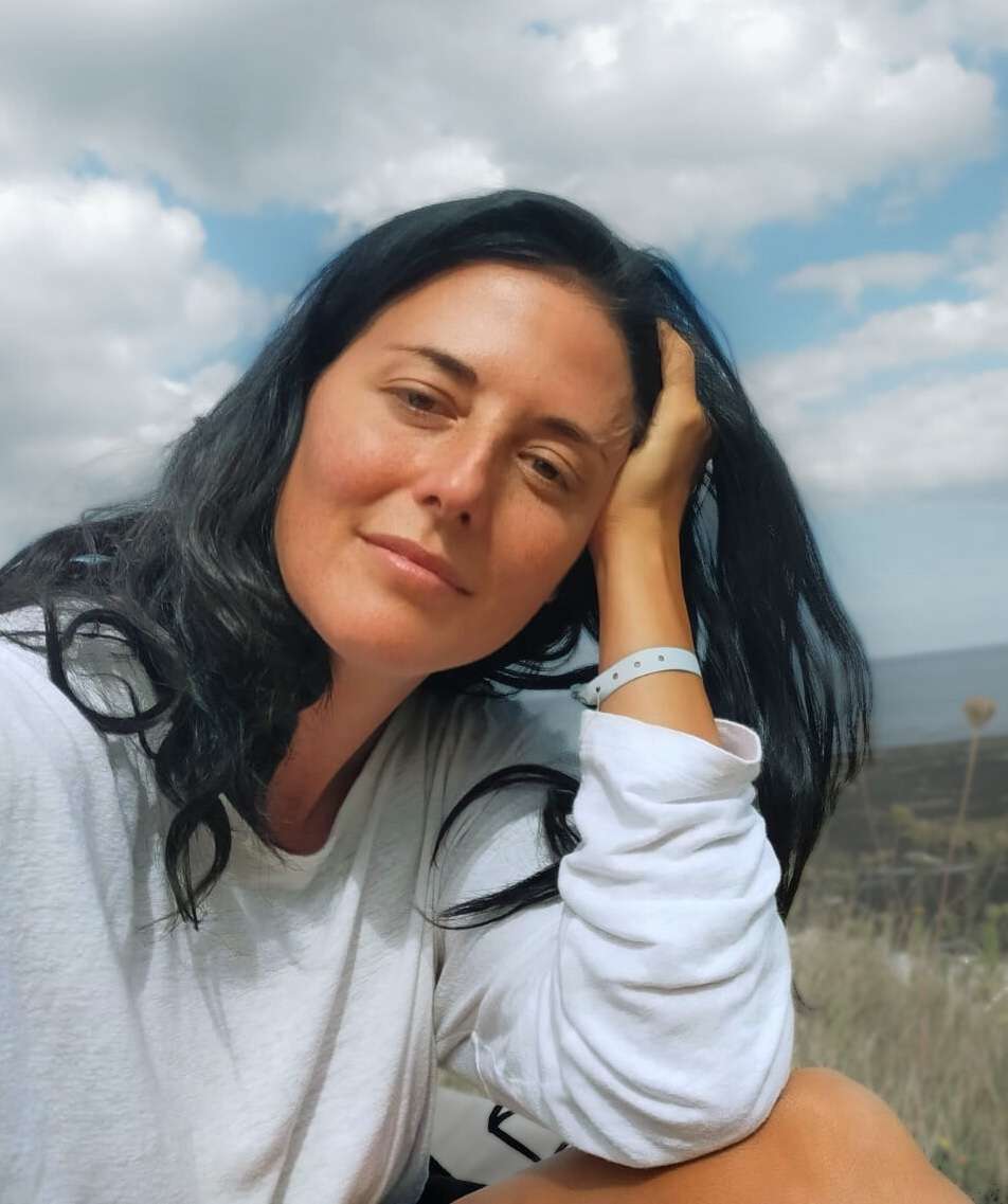 A woman with long, dark hair rests her head on her hand, looking at the camera with a gentle smile. She is outdoors under a partly cloudy sky.