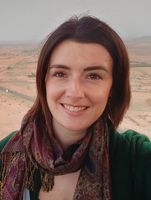 A brown hair and brown eyed woman smiles at camera, with a vast, arid landscape behind her.