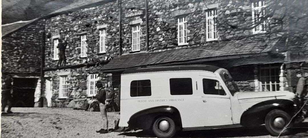 A black and white image from the 1960s of an old ambulance vehicle in front of a slate house.