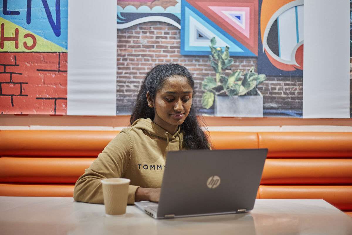 A woman in a hoodie sits at a table, using a laptop, with colorful wall art and a plant in the background.