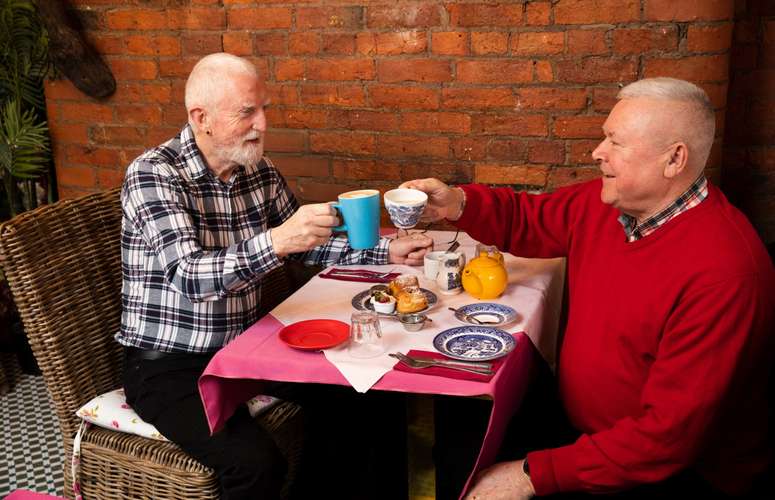 Two men sitting across from each other at a table raising their drinking vessels