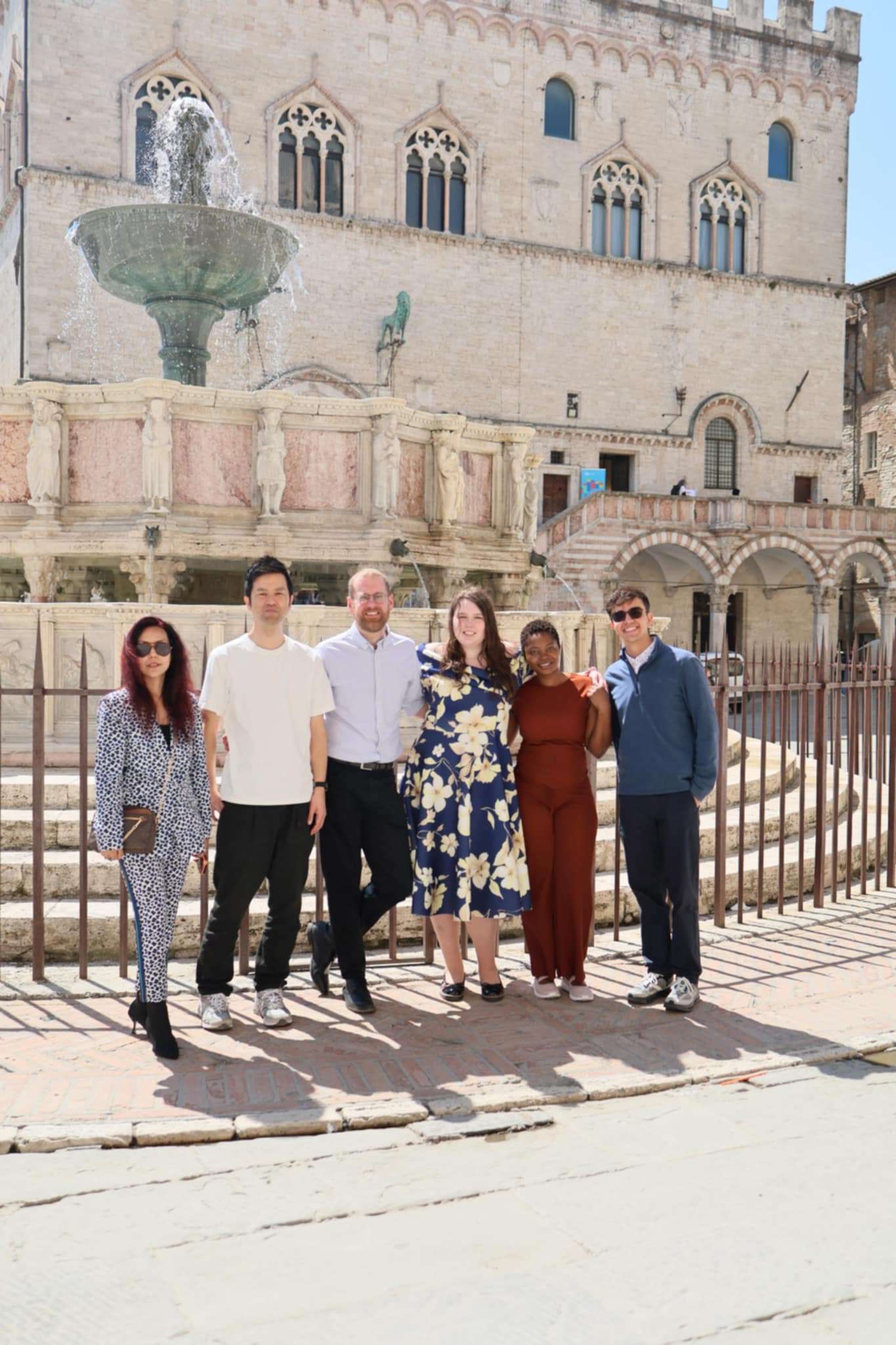 A diverse group of students and staff on a university research trip abroad, posing in front of a historic European fountain on a sunny day.