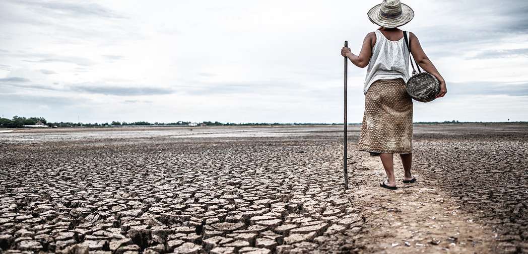 A woman on dry, arid land looking into the distance. She is holding a basket and a cane, and wearing a hat.