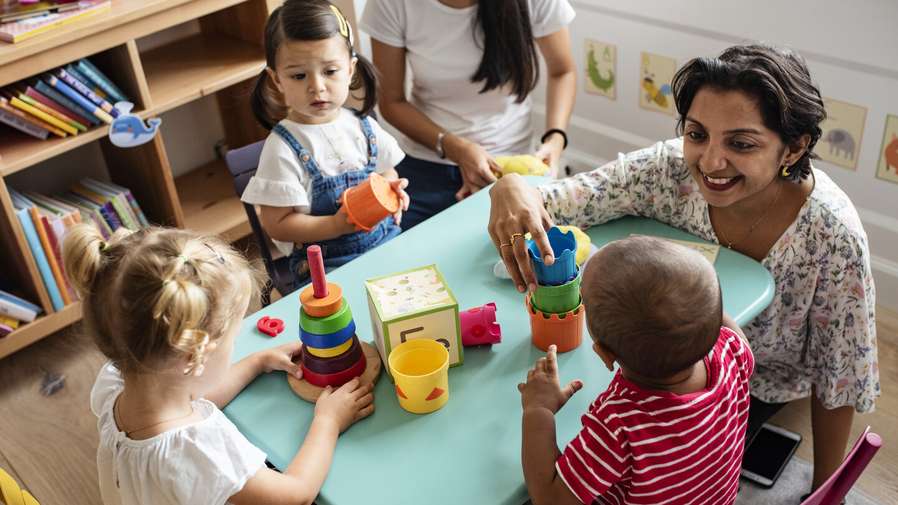 Two adults and three children sit around a table while the children play with toys.