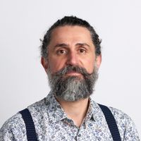 Man with a gray beard and mustache, wearing a floral shirt and suspenders, against a plain white background.
