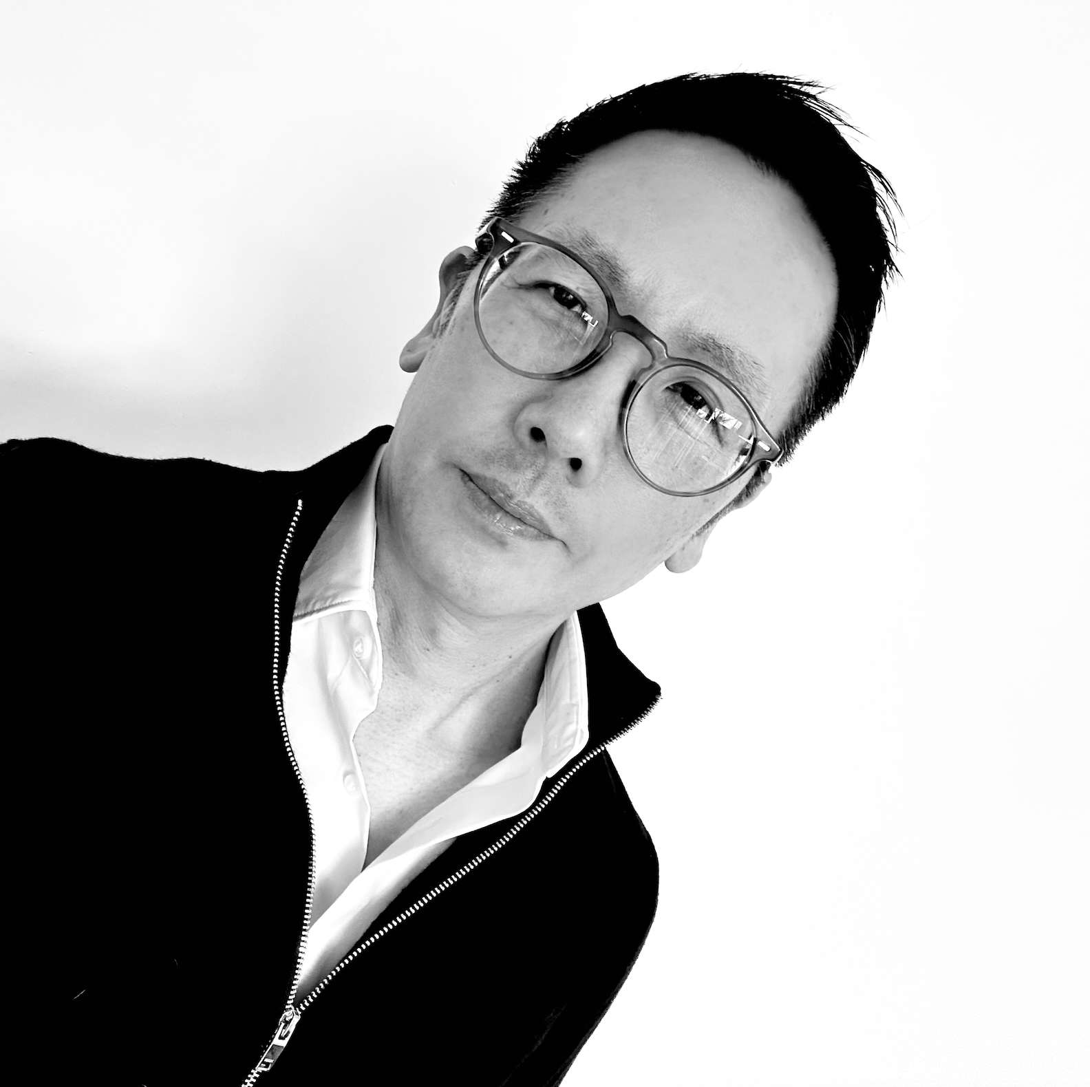 Black and white portrait of a person wearing glasses and a zip-up jacket, looking at the camera against a plain background.