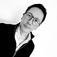 Black and white portrait of a person wearing glasses and a zip-up jacket, looking at the camera against a plain background.