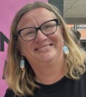 A headshot of a smiling woman with blonde hair, wearing black-rimmed glasses and earrings shaped like ice lollies.
