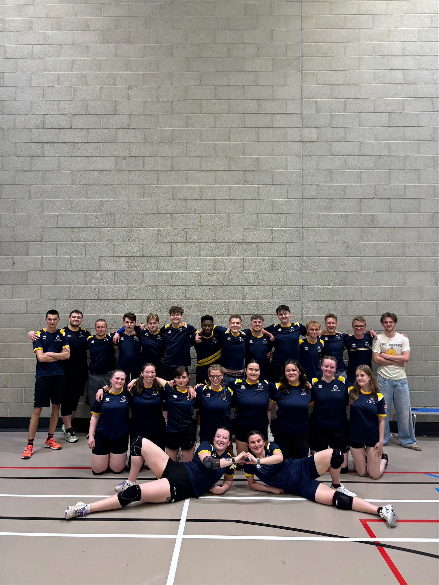 UEA Dodgeball team in navy uniforms posing in a gym setting; two players lie in front forming a heart with their hands.