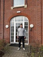 A person stands in front of an arched brick entrance with glass doors, wearing a grey sweater and black trousers, on a cobblestone path.