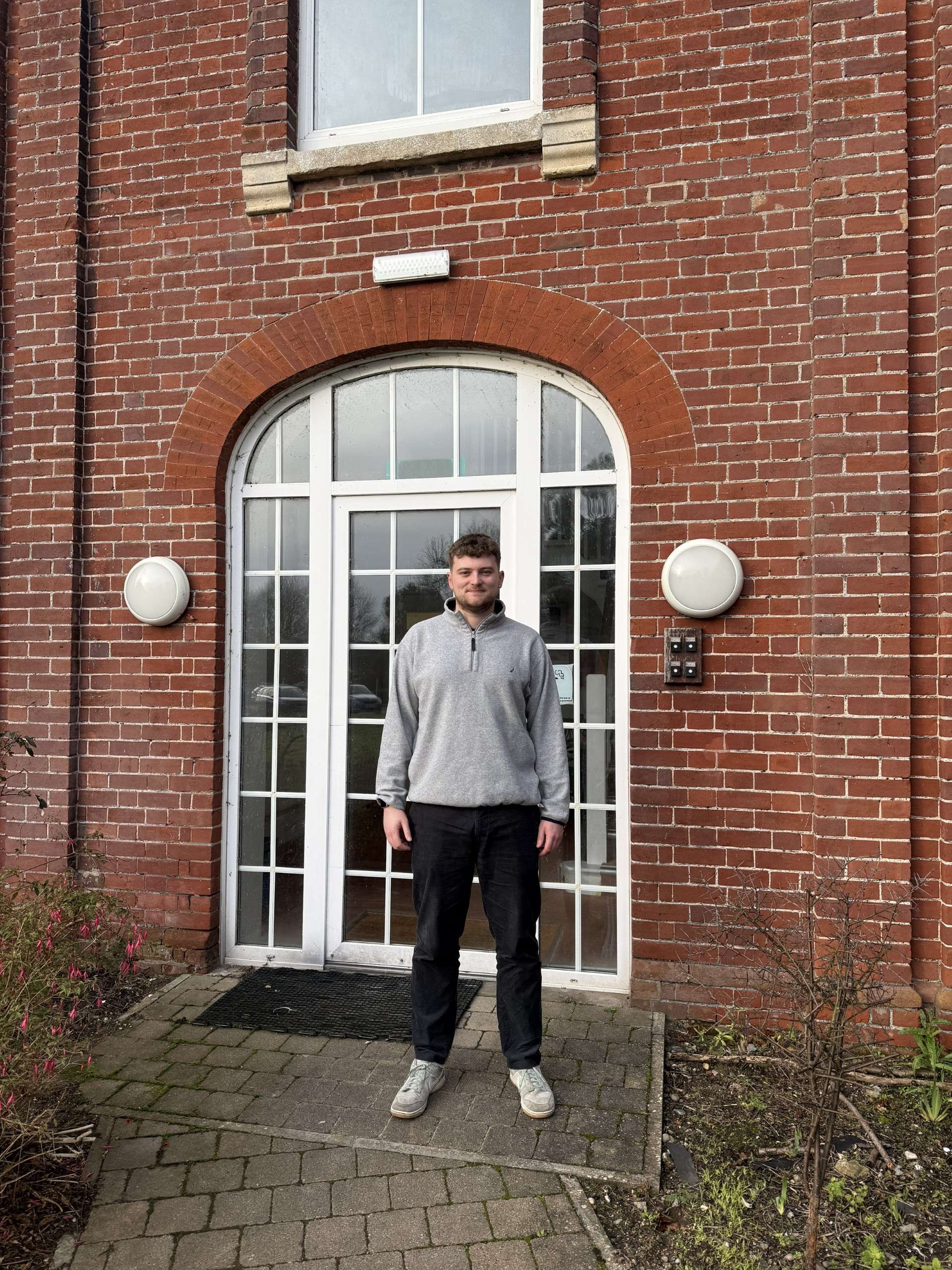 A person stands in front of an arched brick entrance with glass doors, wearing a grey sweater and black trousers, on a cobblestone path.
