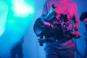 Someone holds a film camera. The background is lit in pinks and blues.