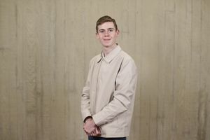 Young man in a beige jacket poses in front of a textured wooden background, smiling confidently at the camera.