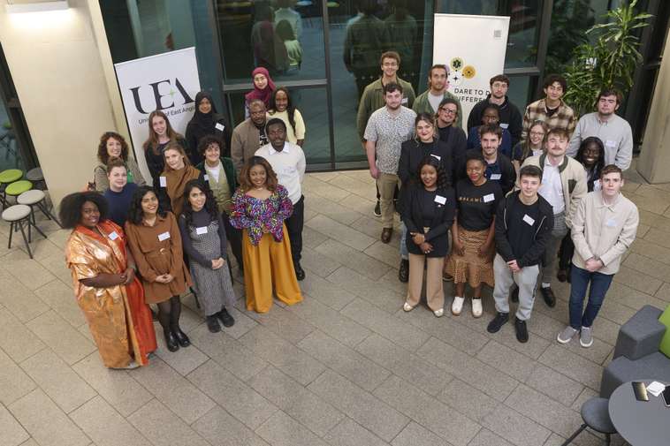 A diverse group of people posing together indoors, with banners and a plant in the background.