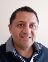 Smiling man with short dark hair wearing a navy shirt with a gray collar against a plain background.