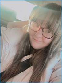 Holly with long hair and glasses sitting in a car, wearing a pink coat. Sunlight filters in from the window, creating a soft glow.