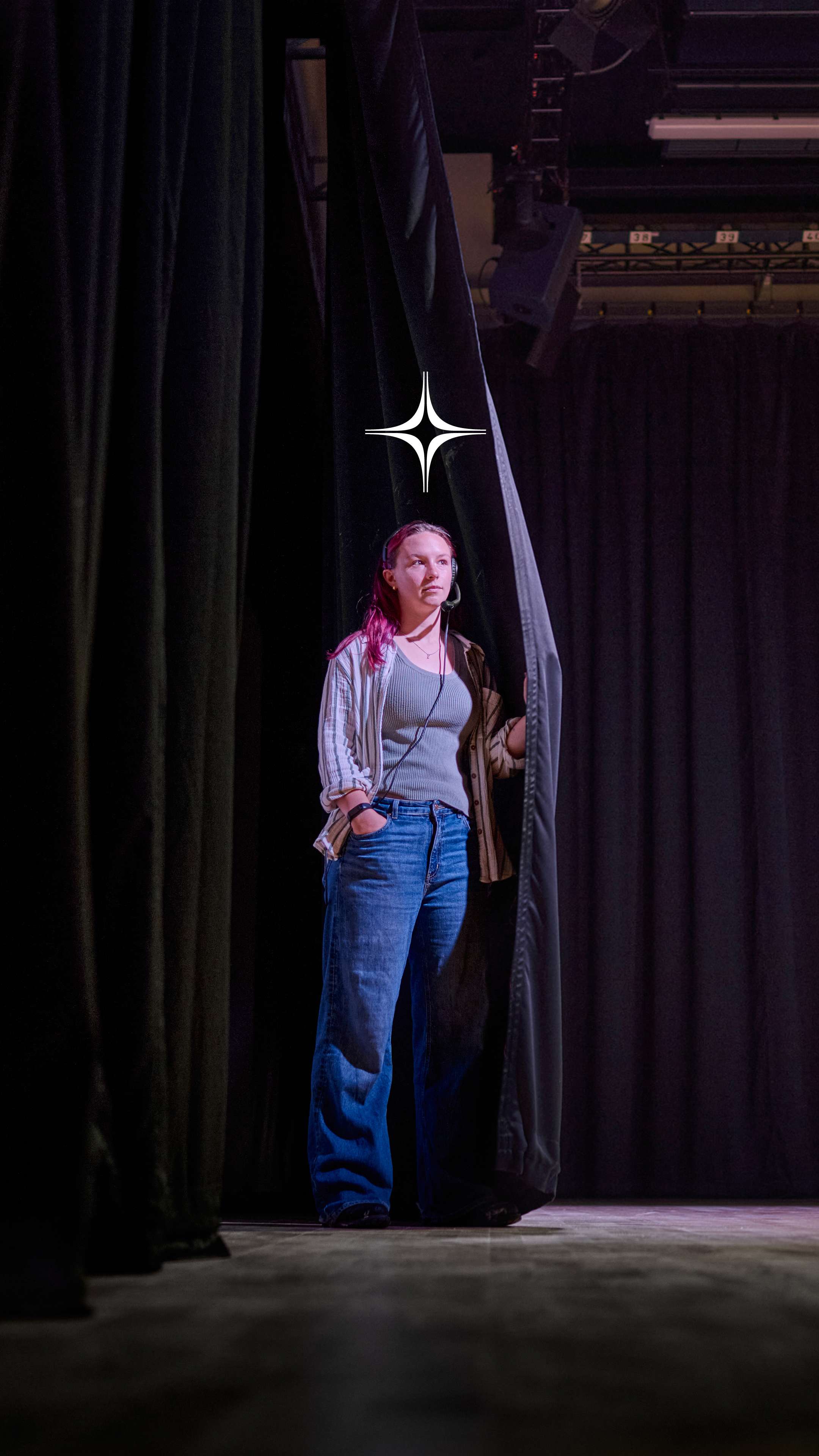 A student stands in the wings of a stage. They have a headset on.
