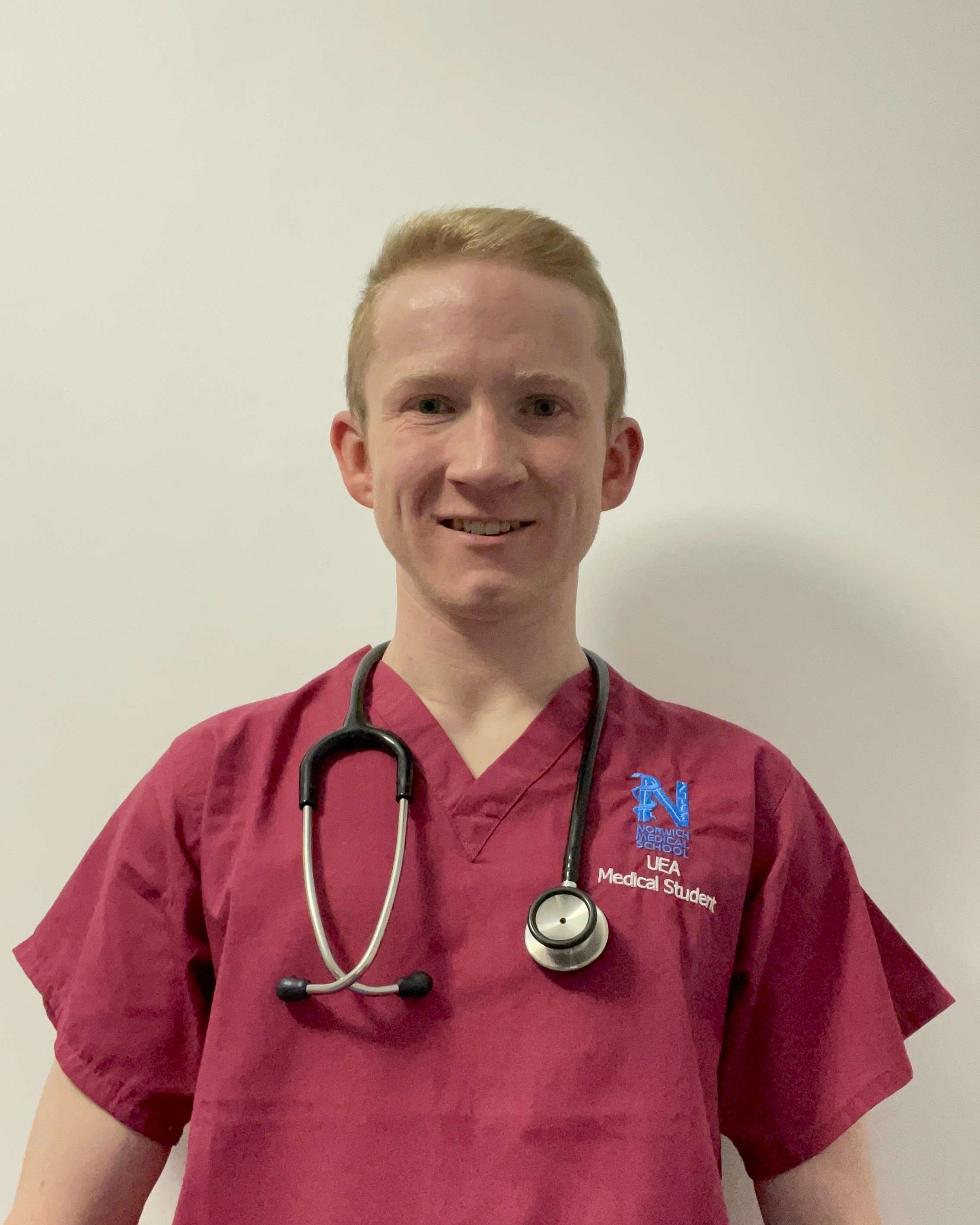 Alex Gibbs wearing maroon scrubs with a stethoscope around his neck, smiling at the camera and standing in front of a cream-colour wall.