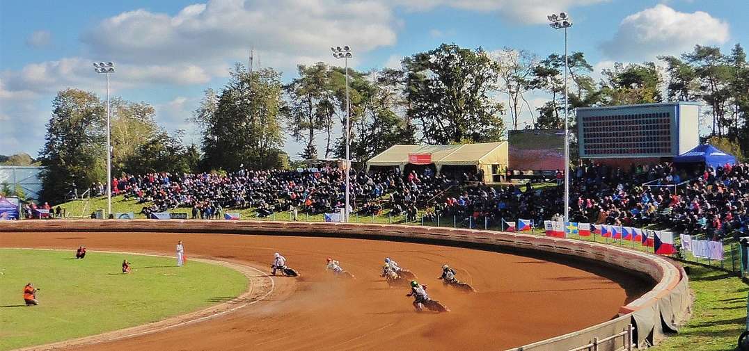Motorcycle racers speed around a dirt track, with spectators in stands under a partly cloudy sky and surrounded by trees.