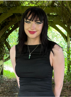 A smiling postgraduate student with black hair, blakck winged eyeliner and a green crystal necklace stands under a leafy garden archway