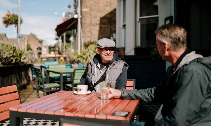 Two men sat outside at a table, with a glass of water and a cup of tea between them