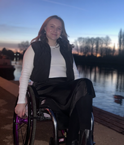 Holly in a wheelchair smiles by a riverside at sunset, wearing a white shirt, black vest, and skirt. Silhouetted trees line the horizon.