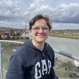 Isaac in a GAP hoodie smiles on a balcony overlooking a river and houses, under a cloudy sky.