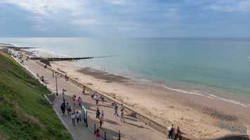 Image of a beach and the ocean on a sunny day, with a green slope on the left, a boardwalk with multiple people, then the beach and the ocean on the right with a groyne in the distance