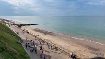 Image of a beach and the ocean on a sunny day, with a green slope on the left, a boardwalk with multiple people, then the beach and the ocean on the right with a groyne in the distance