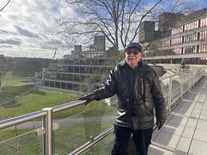 Ian stands smiling on the walkway on a sunny day with the Zigs behind him wearing a leather jacket and gloves, sunglasses and a black cap.