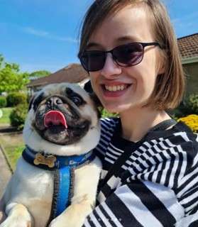 Emma smiling in sunglasses holding a happy pug with its tongue out, outside on a sunny day.