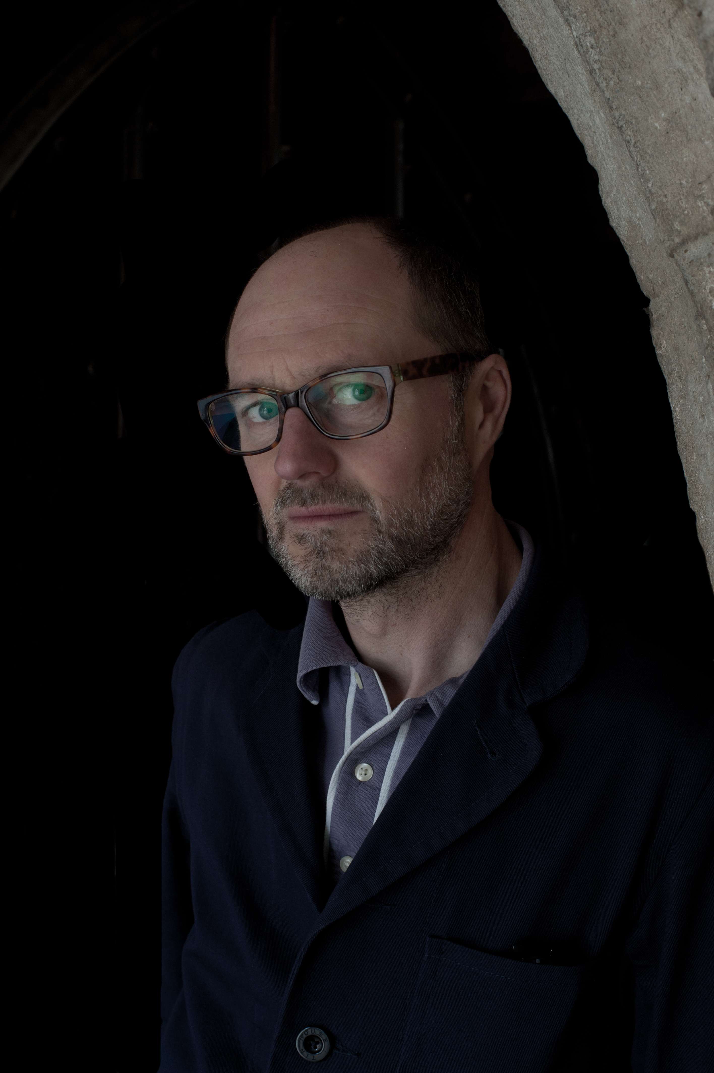 A man with glasses in a dark door way looks at the camera.