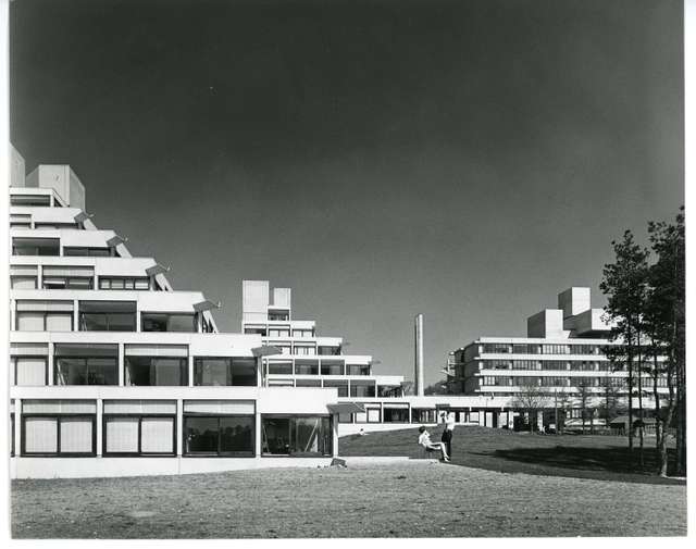 Black and White image of the Ziggurats on a sunny day with a woman sat outside sat on a chair talking to a man.