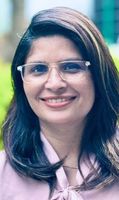 A woman with long dark hair and glasses smiles, wearing a light pink blouse, in an outdoor setting with greenery in the background.