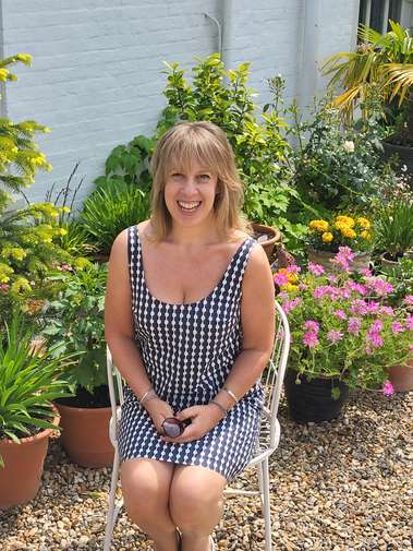 A female academic with blonde hair smiles warmly, seated in a sunny garden surrounded by plants
