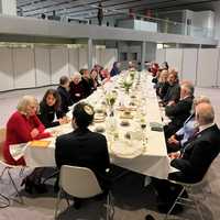 A large group of people sit around a long, elegantly set dining table in a modern room, engaged in conversation.