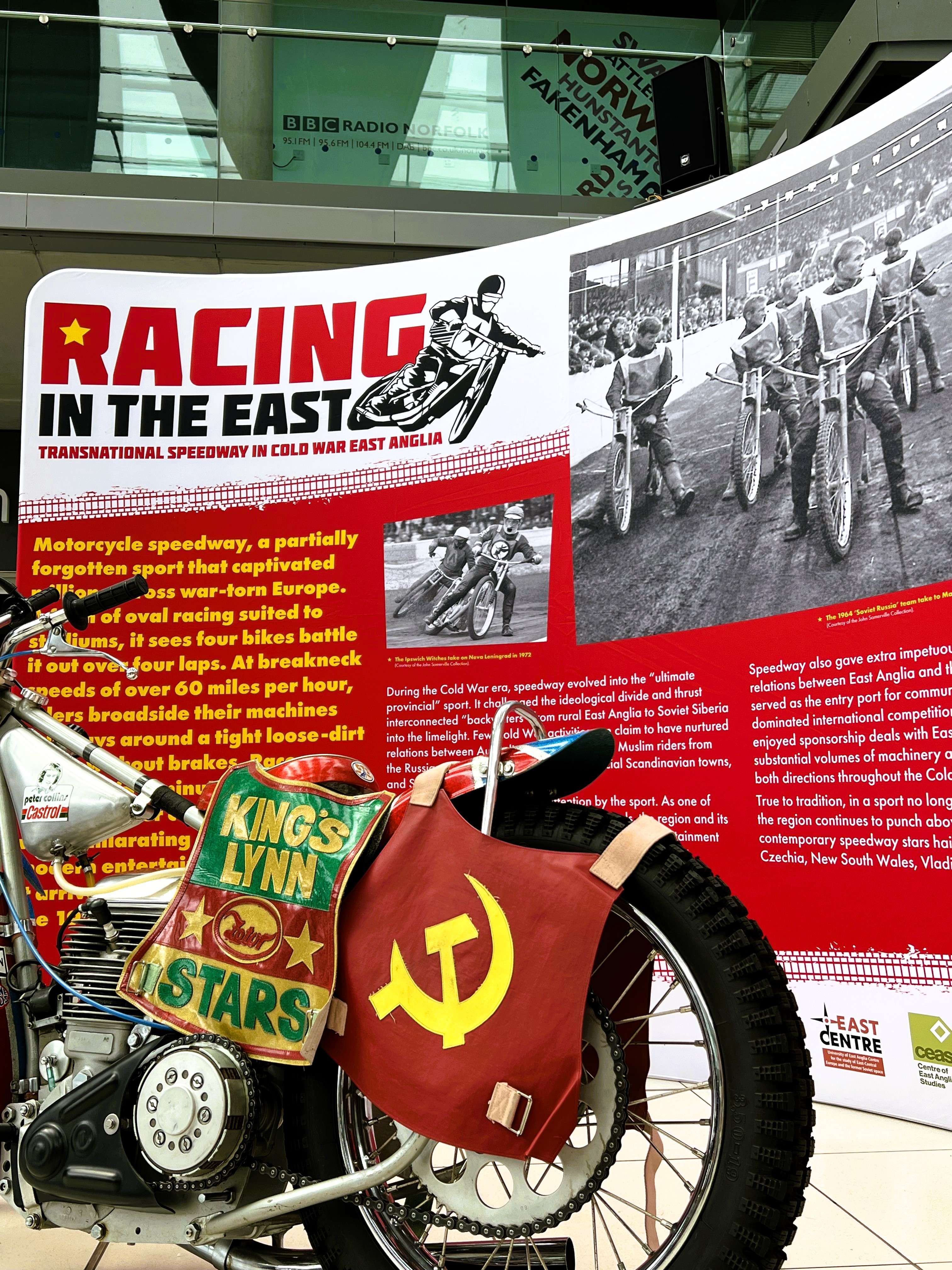 Motorcycle display with racing gear, Soviet flag, and a backdrop detailing the history of speedway racing in Cold War East Anglia.