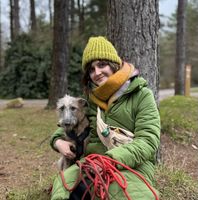 Woman in a green coat and hat sits by a tree with a dog, holding a red leash. They are outdoors in a wooded area.