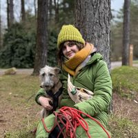 Woman in a green coat and hat sits by a tree with a dog, holding a red leash. They are outdoors in a wooded area.