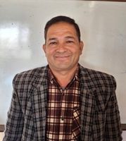 Smiling man in a plaid shirt and checkered jacket stands in front of a whiteboard.