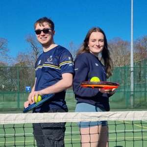 Two UEA Tennis members standing back-to-back behind a tennis net, each holding a racket with tennis balls, smiling on an outdoor court under blue sky.